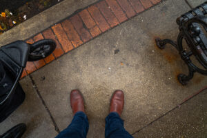 Looking down at the feet of a man wearing jeans and brown leather shoes standing on the concrete and brick paver sidewalk 